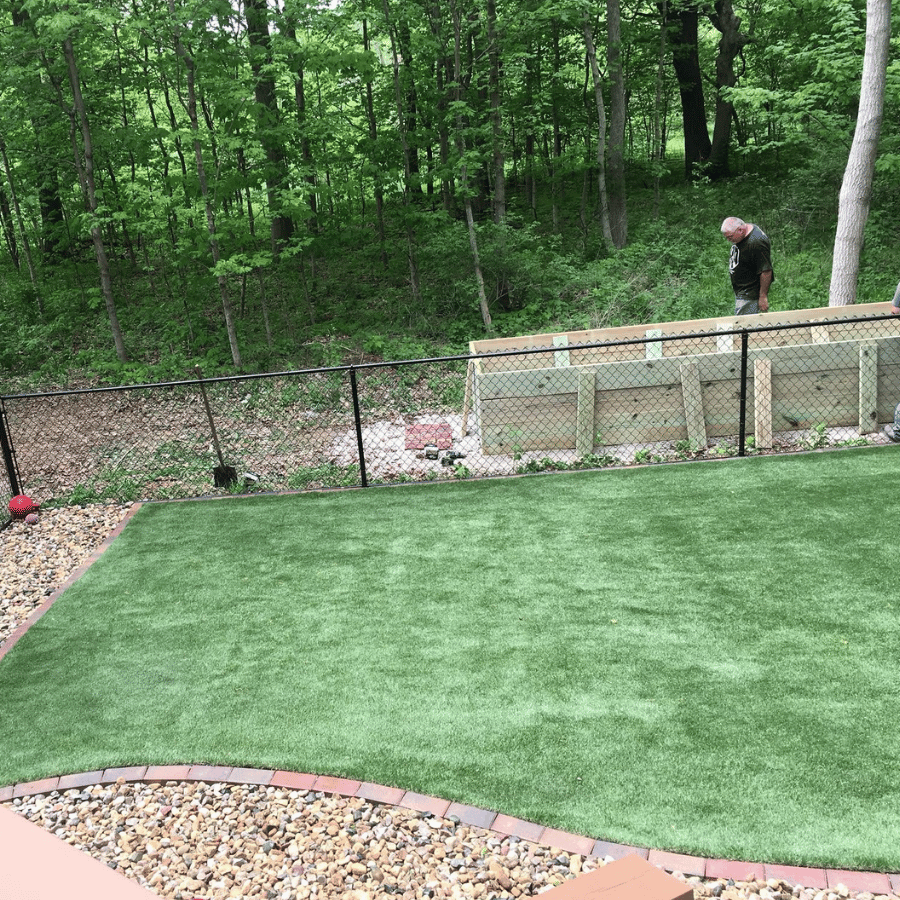 Man stands by a fence in a backyard with artificial grass, surrounded by trees. A wooden structure is visible in the background.