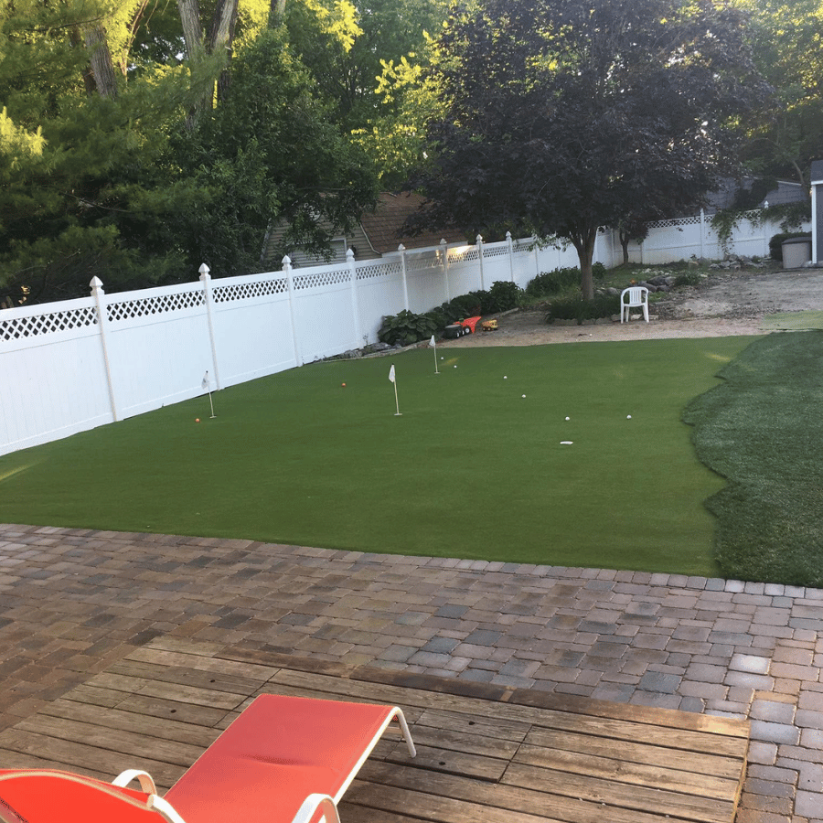 Backyard with a putting green, white fence, and trees. A red lounge chair sits on the patio, creating a relaxing atmosphere.