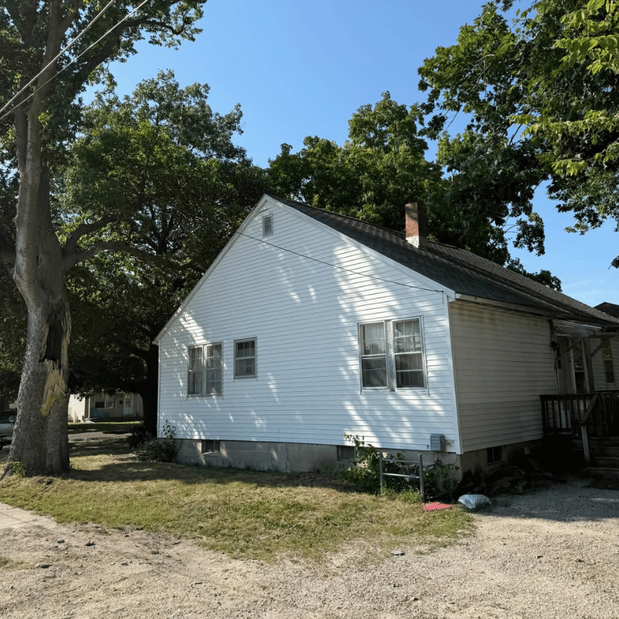 A small white house with a gabled roof sits in sunlight, surrounded by trees and a gravel path, under a clear blue sky.