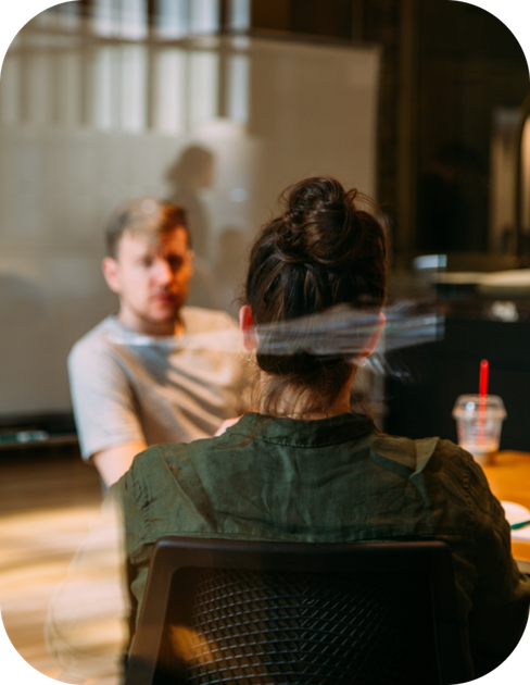 Two people talking across a table in a glass-walled office, with drinks and warm light.