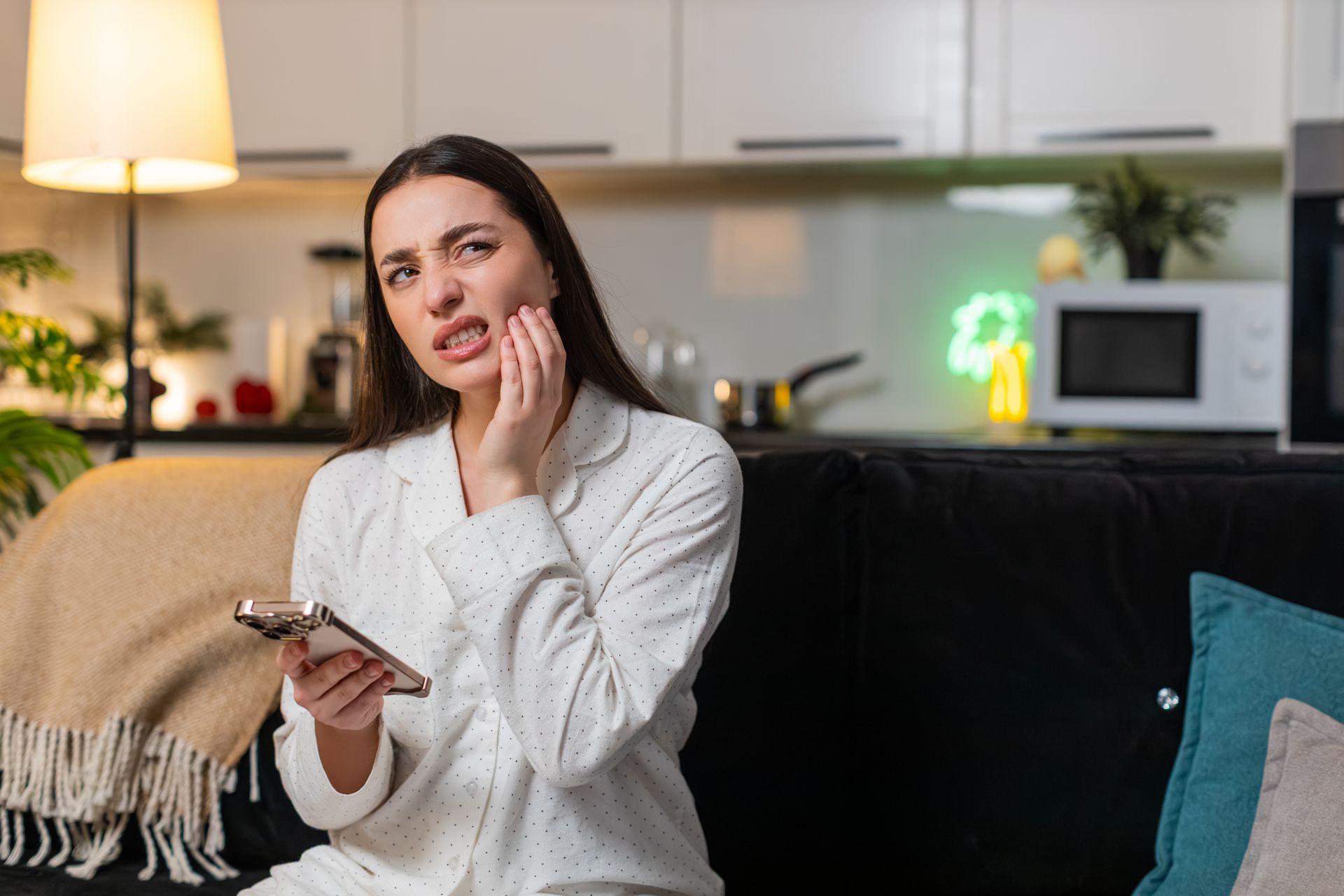 A woman at home, holding her cheek with tooth pain, uses her smartphone to find a dentist.