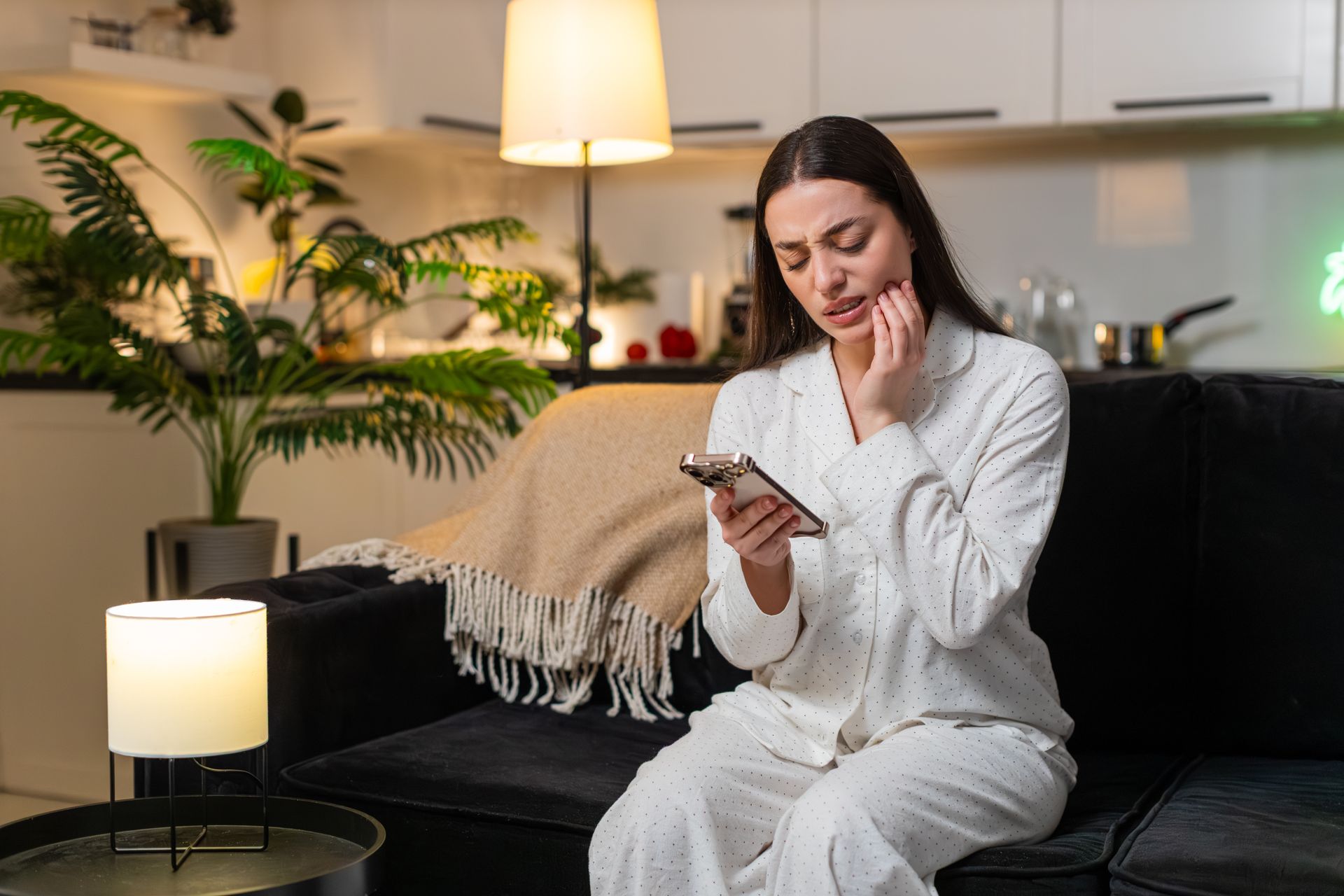 A woman at home, holding her cheek with tooth pain, uses her smartphone to find a dentist.