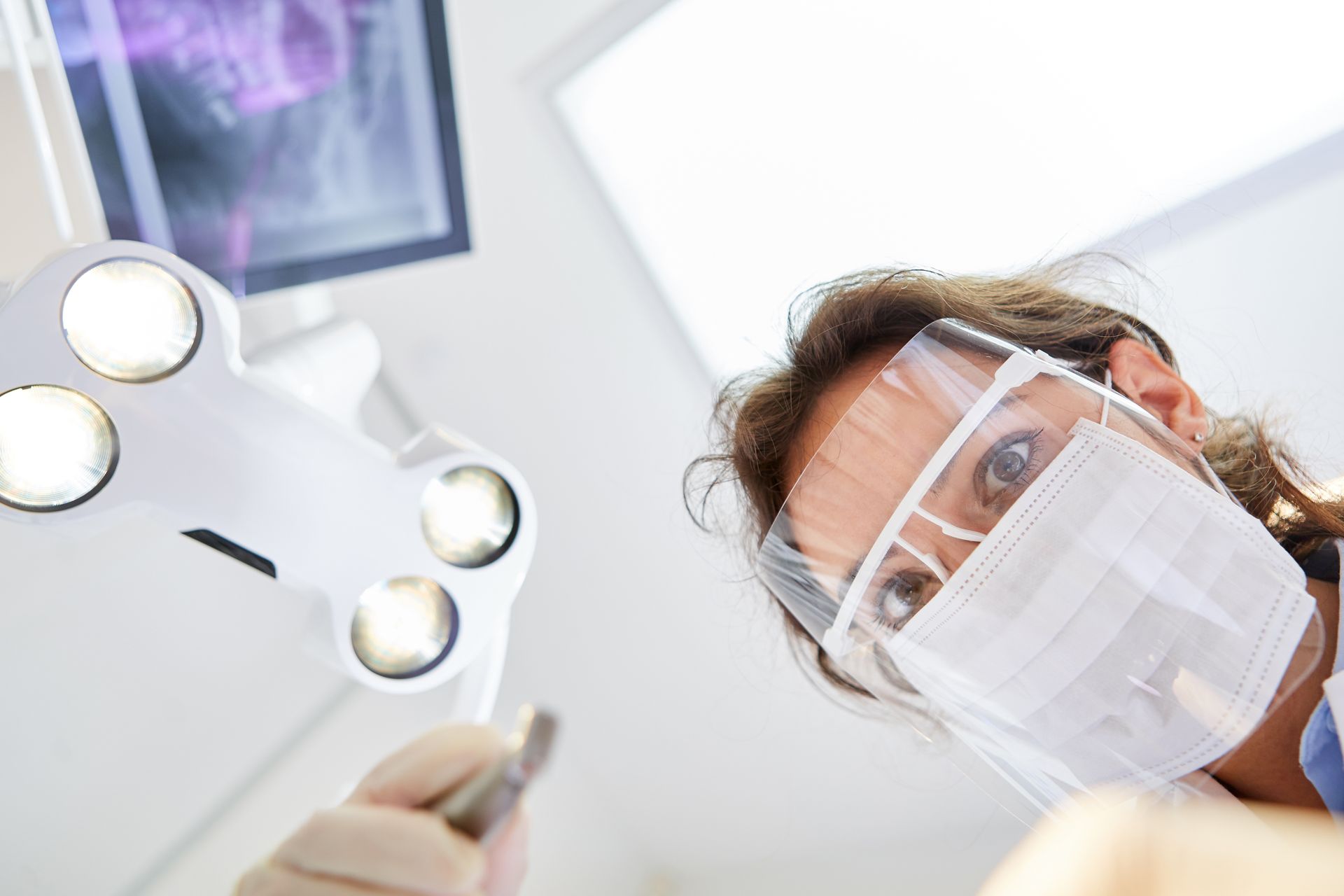 A dentist looking down at the camera with a surgical mask and visor.