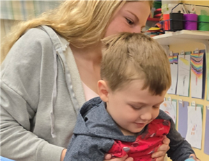 Woman in gray hoodie holding a smiling child in a classroom, with colorful artwork in the background