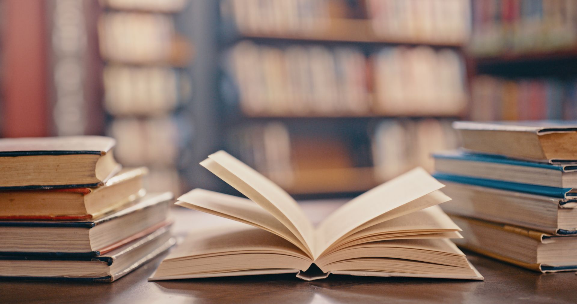 Open book on table, stacks of books on either side, bookshelves in the background.