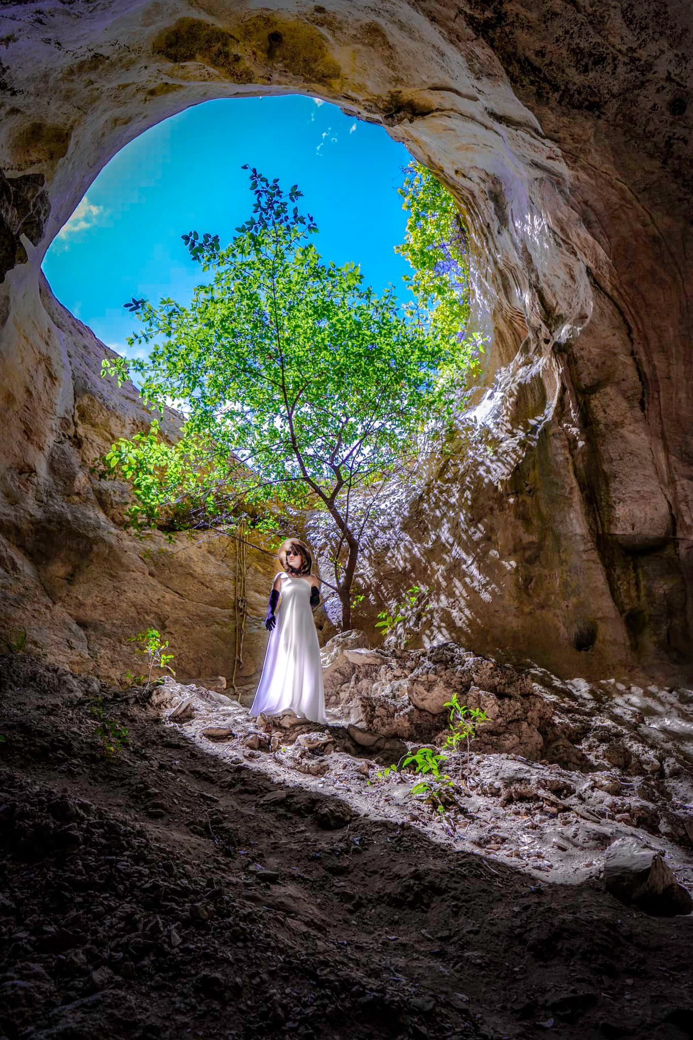 A woman in a white dress is standing in a cave with a tree in the background.