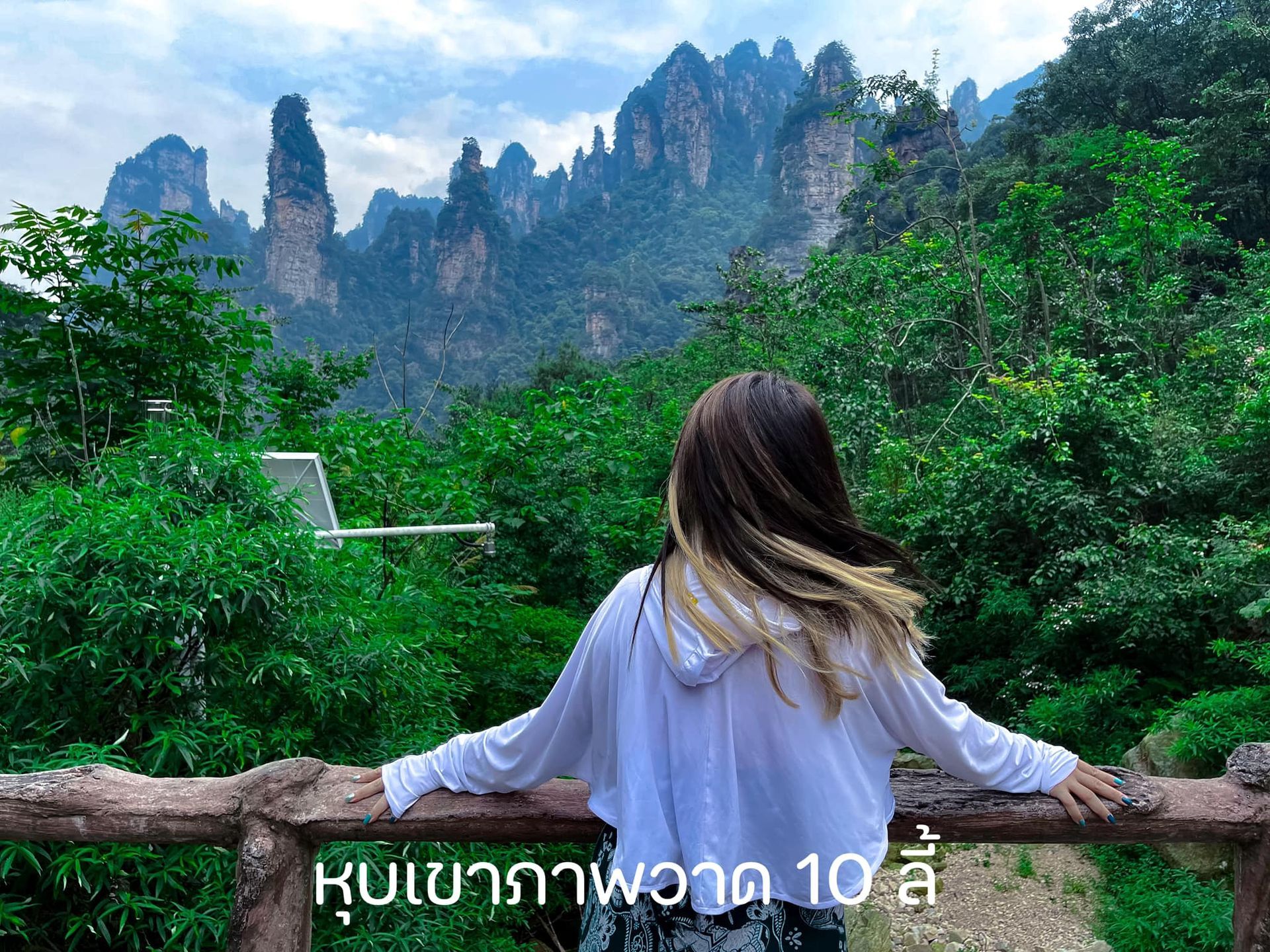 A woman is standing on a wooden railing looking at a mountain range.