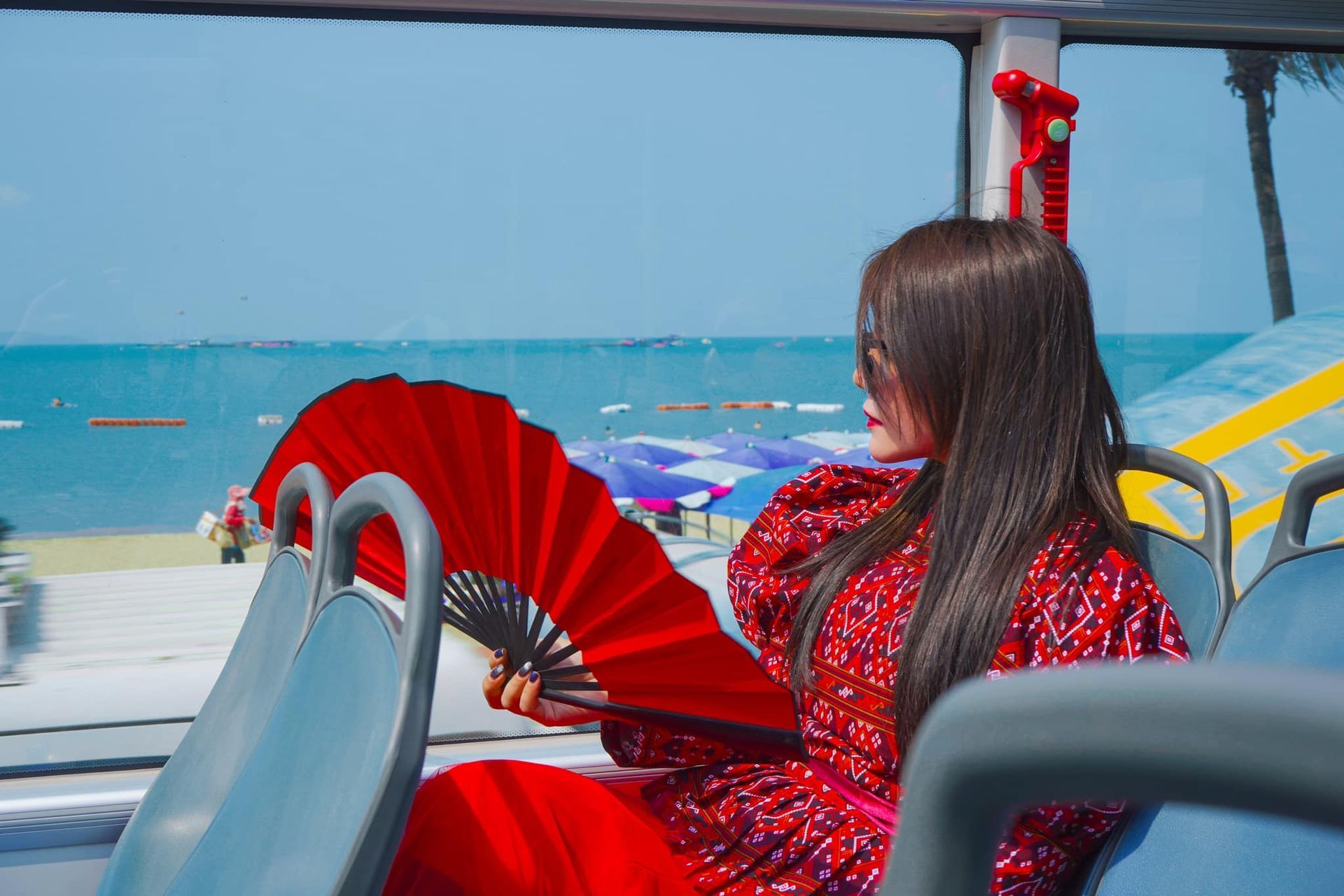 A woman is sitting on a bus holding a red fan.