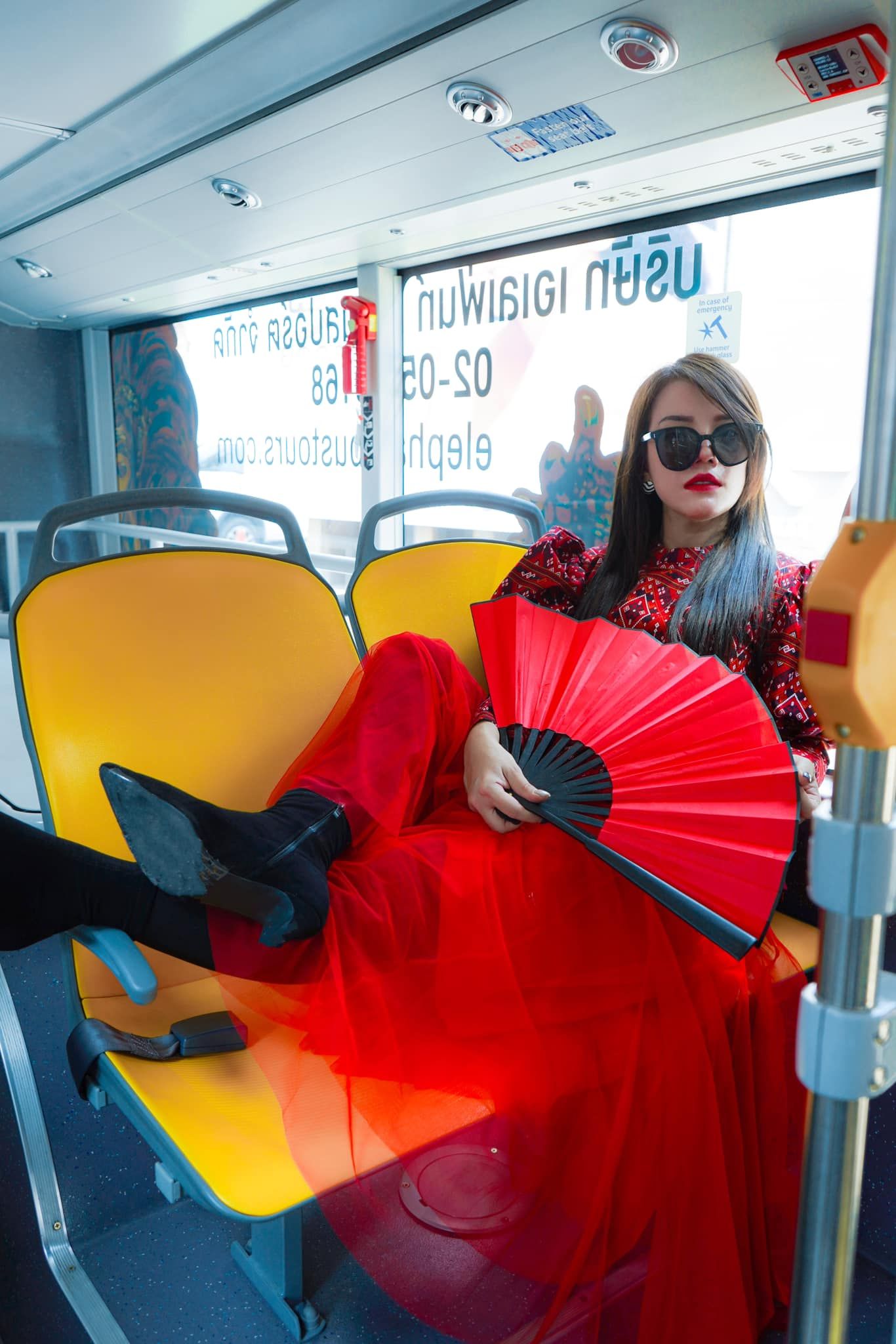 A woman in a red dress is sitting on a bus holding a red fan.