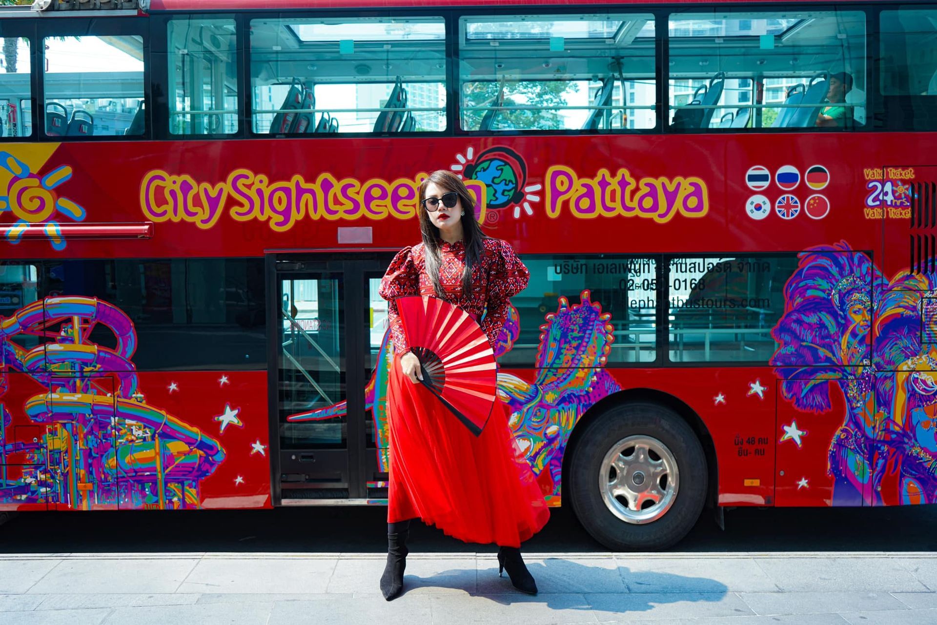 A woman in a red dress is holding a fan in front of a red double decker bus.