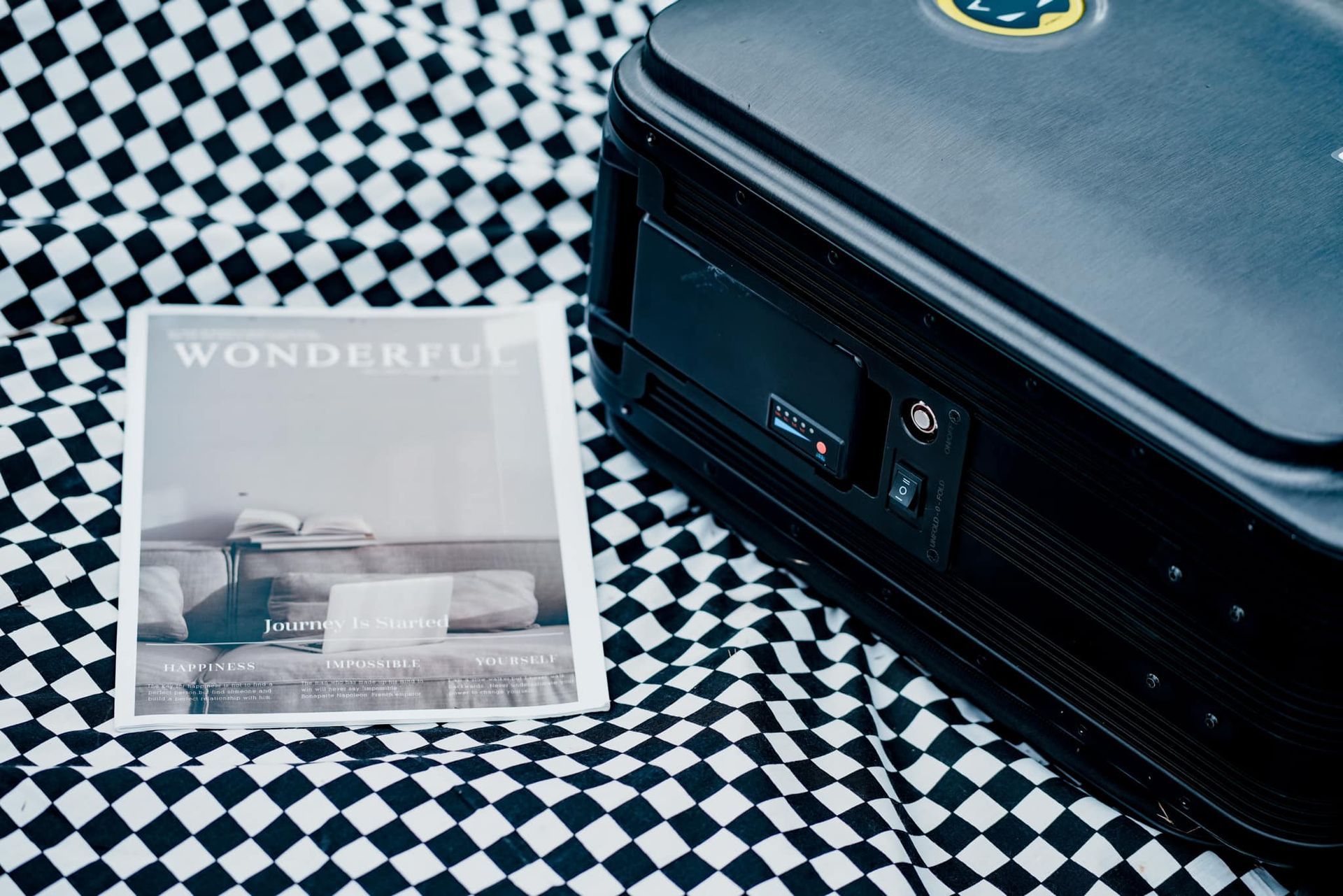 A black suitcase is sitting on a checkered table next to a magazine.