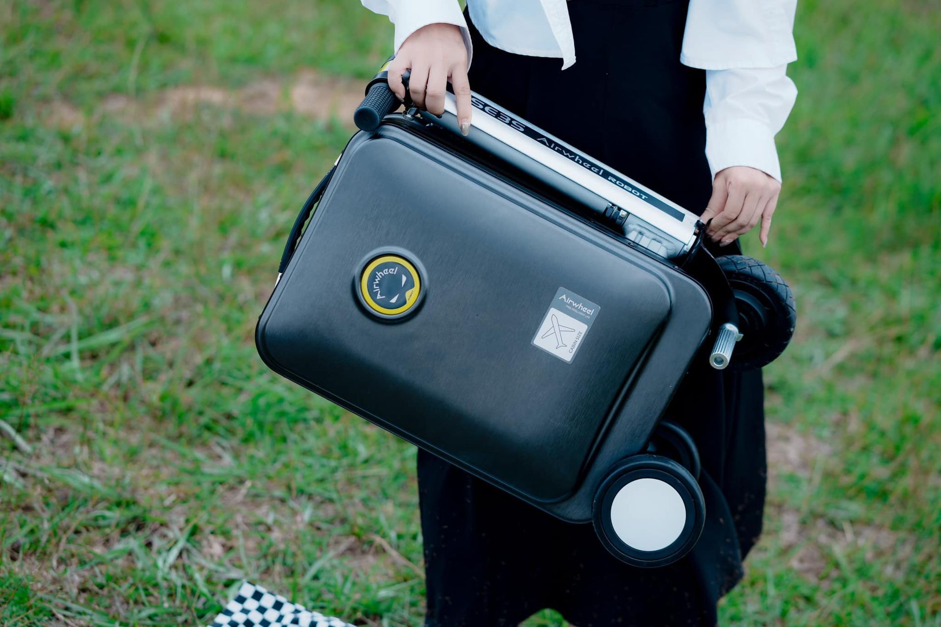 A woman is holding a black suitcase in a field.