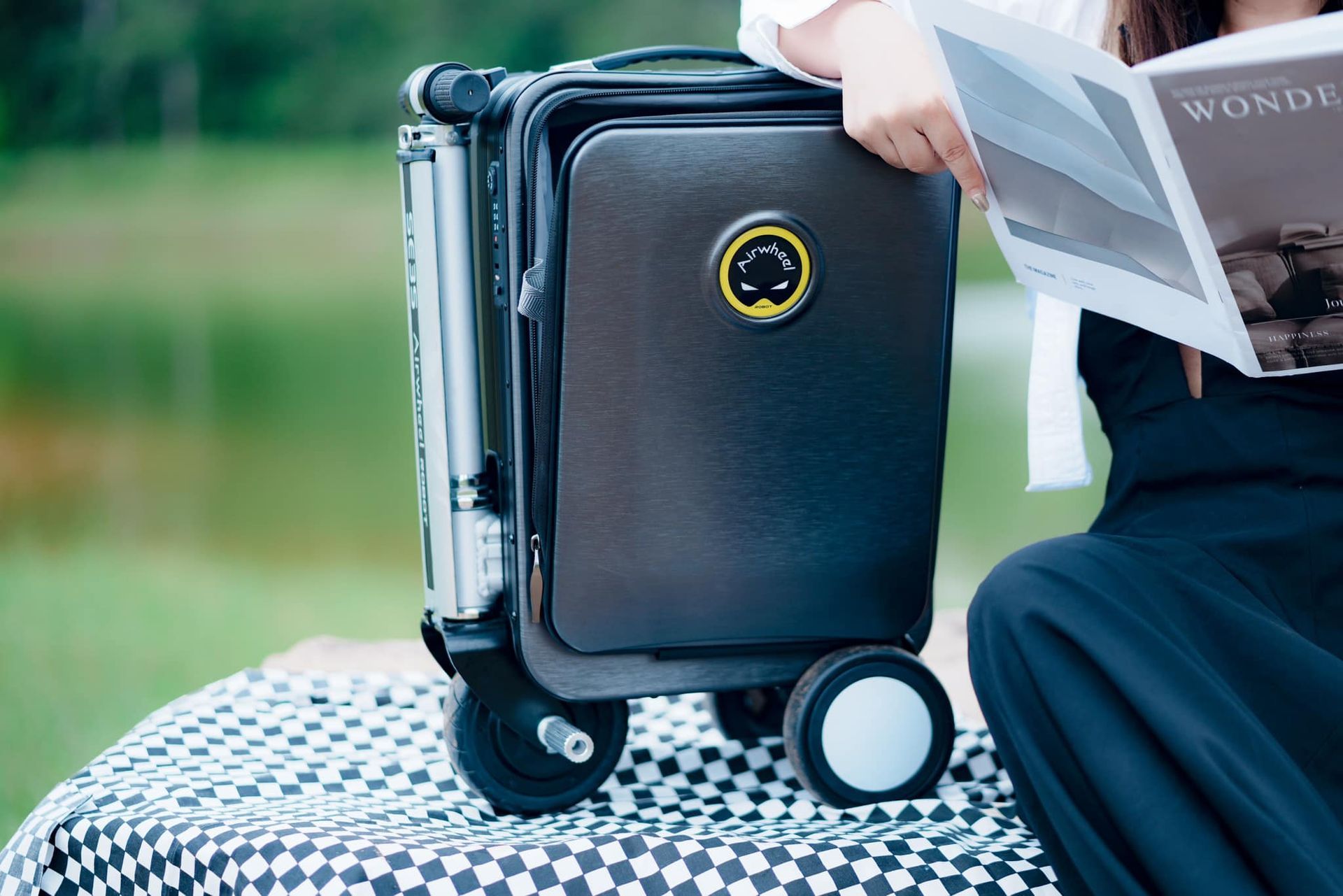 A woman is sitting on a blanket with a suitcase and reading a magazine.