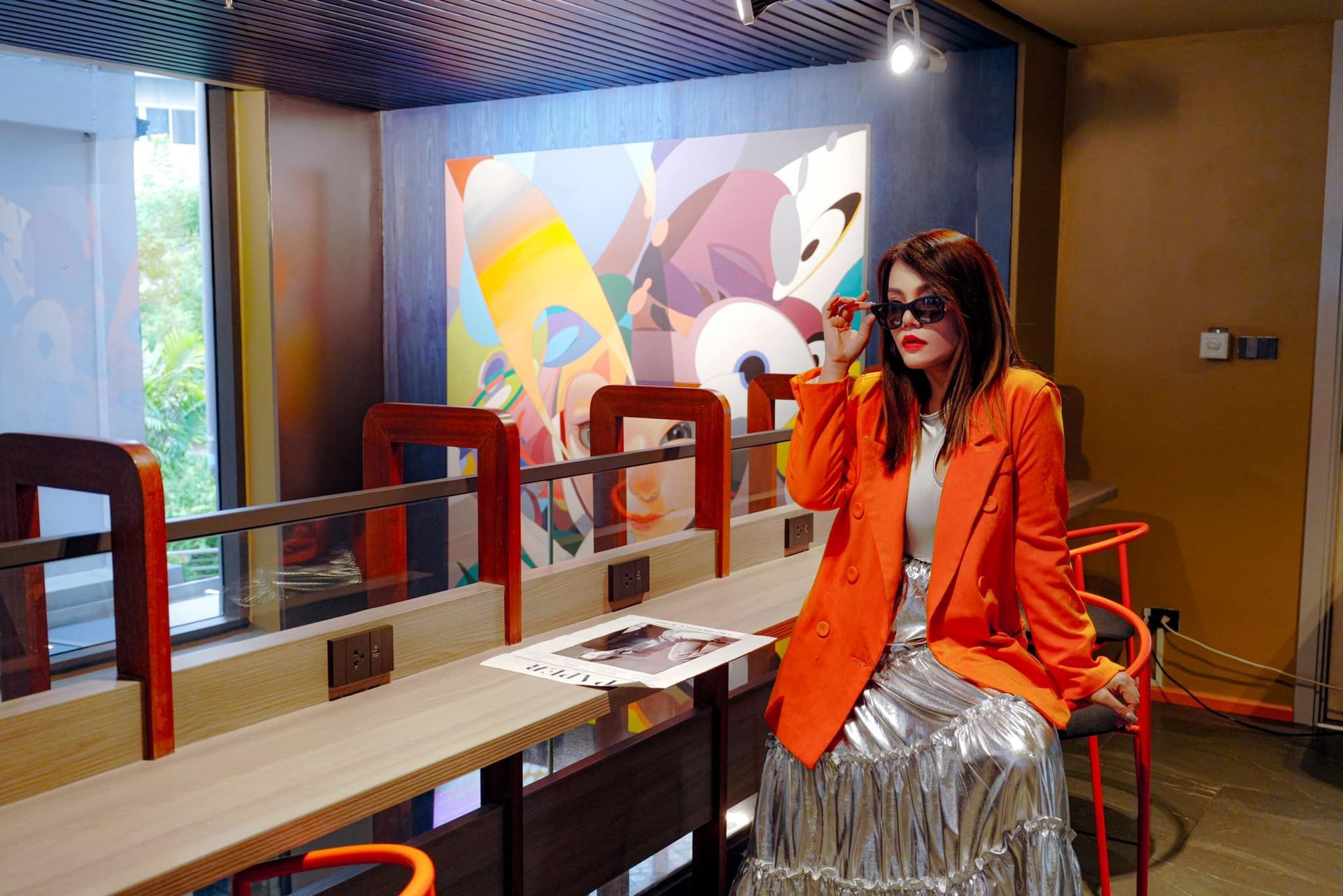 A woman in an orange jacket is sitting at a table in front of a painting.
