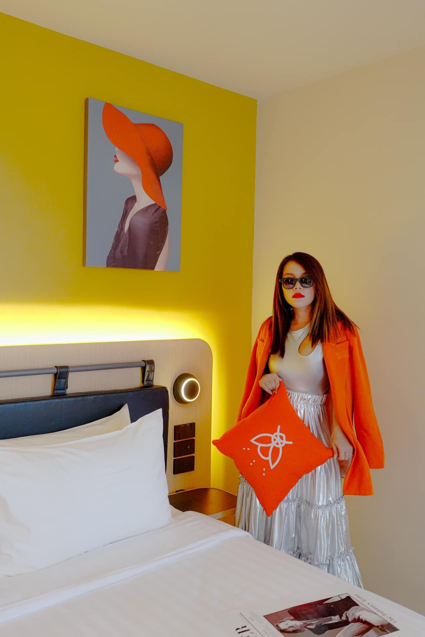 A woman is standing next to a bed in a hotel room holding an orange pillow.