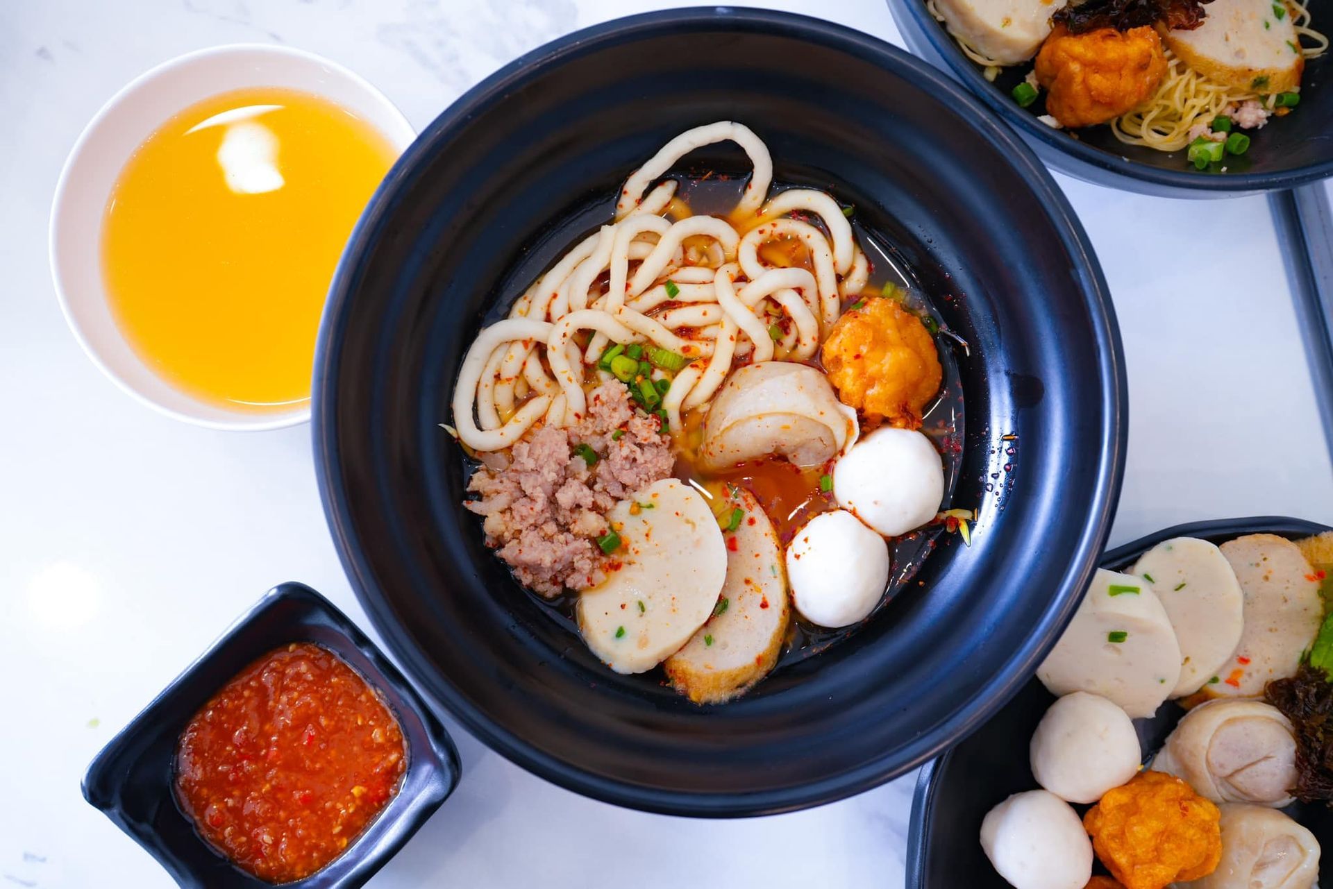 A bowl of noodles with fishballs and sauce on a table.