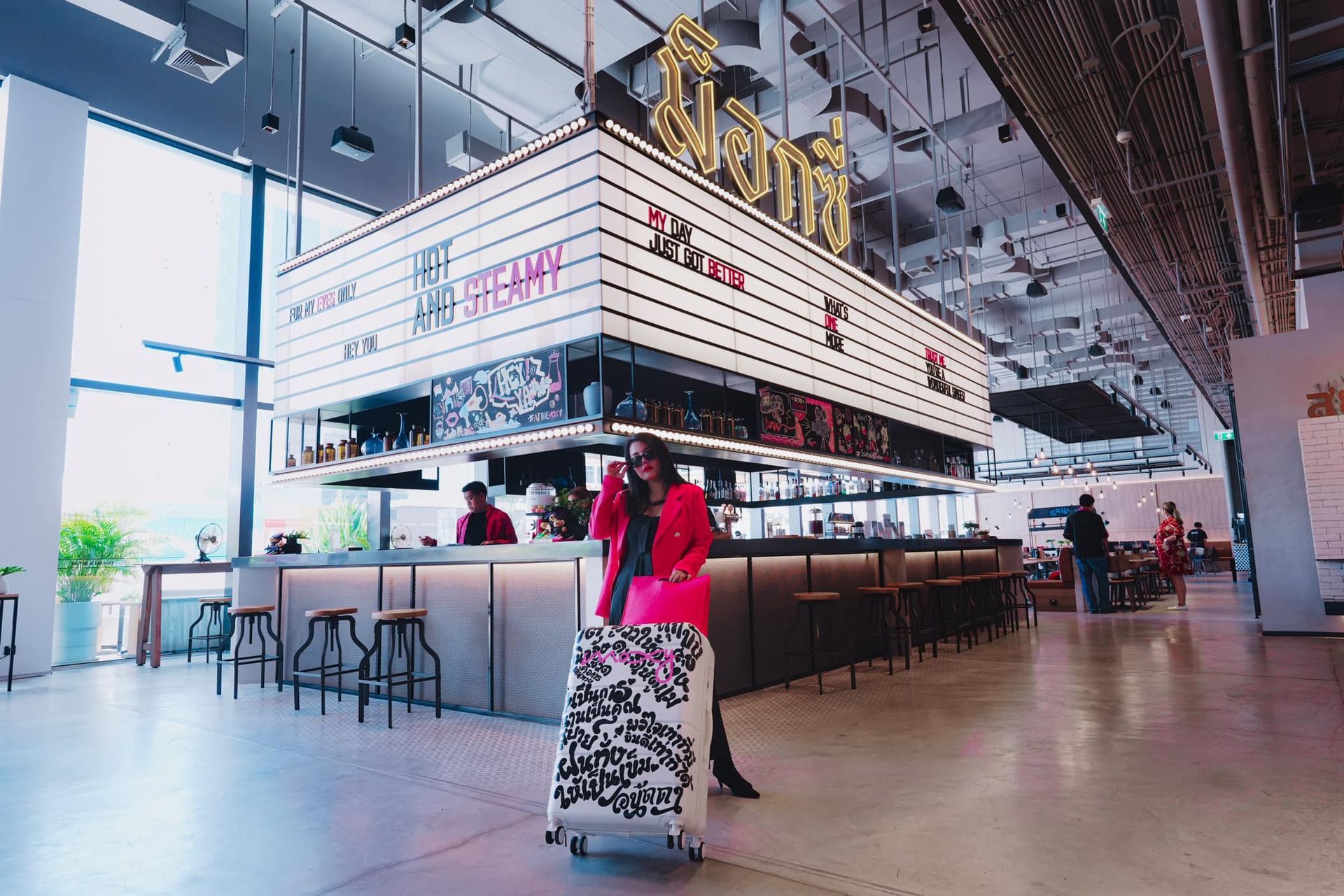 A woman is pushing a suitcase in front of a sign that says in Moxy Bangkok Ratchaprasong hotel.