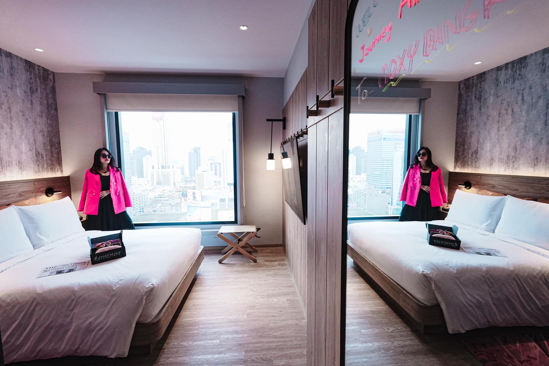 A woman in a pink jacket is standing in a hotel room next to two beds.