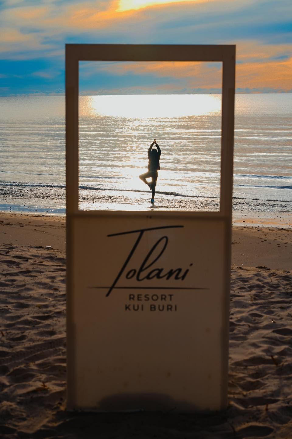 A woman is doing yoga on the beach in front of a tolani resort  Kuiburi sign
