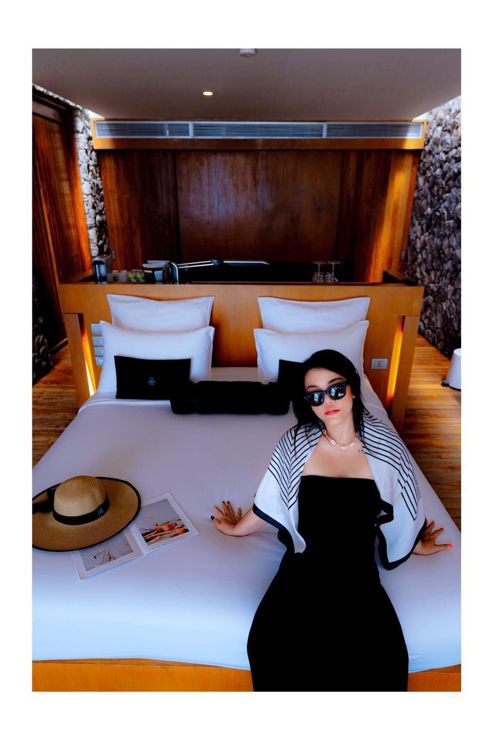 A woman in a black dress is sitting on a bed in a hotel room.