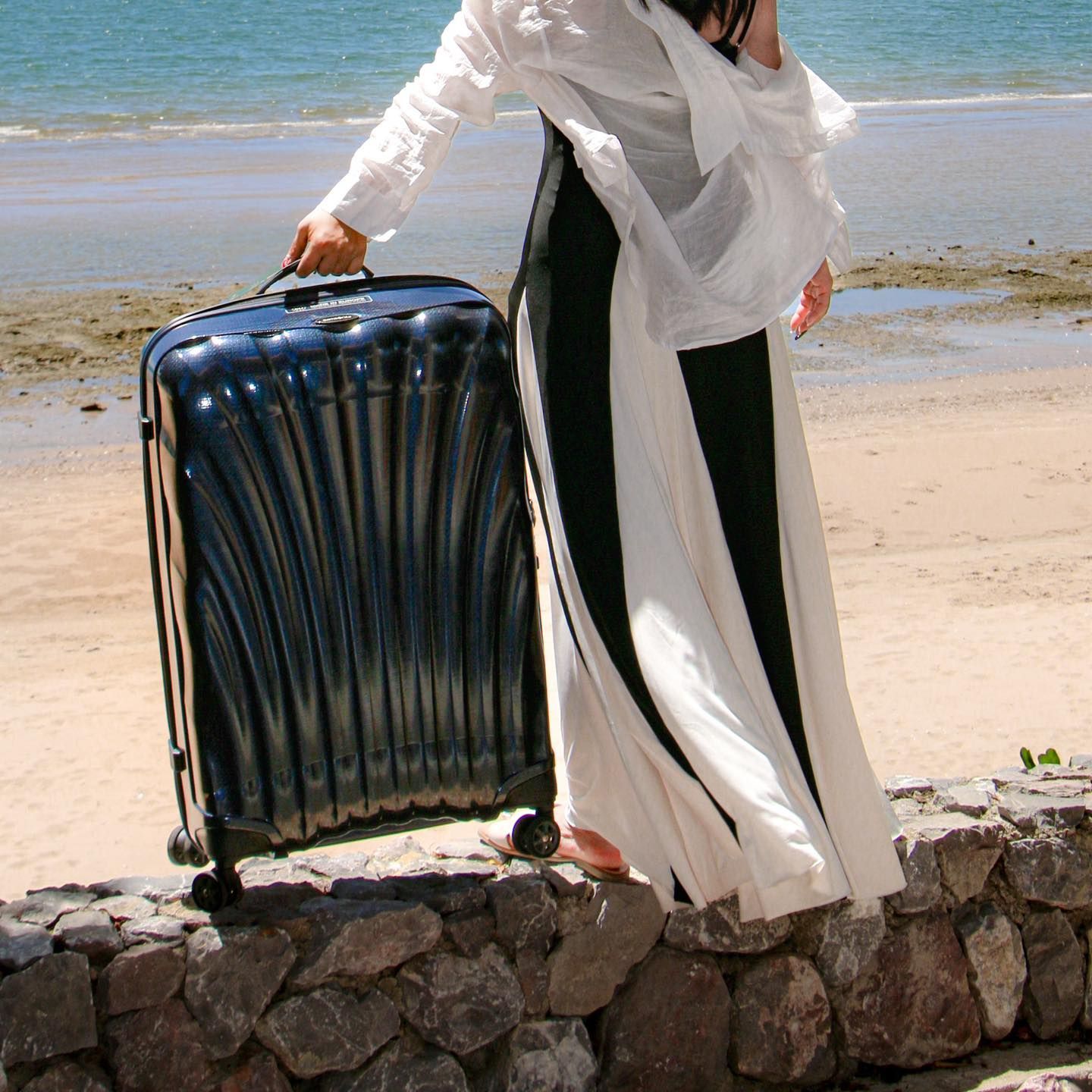 A woman in a white dress is holding a suitcase on the beach