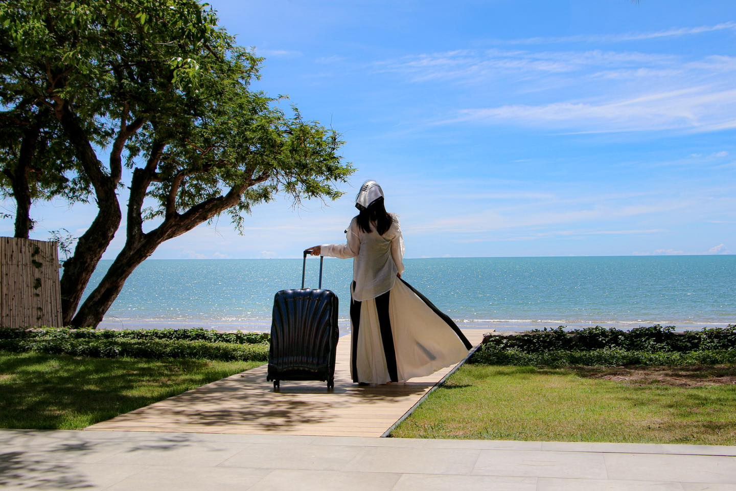 A woman is standing next to the samsonite suitcase on a sidewalk looking at the ocean.