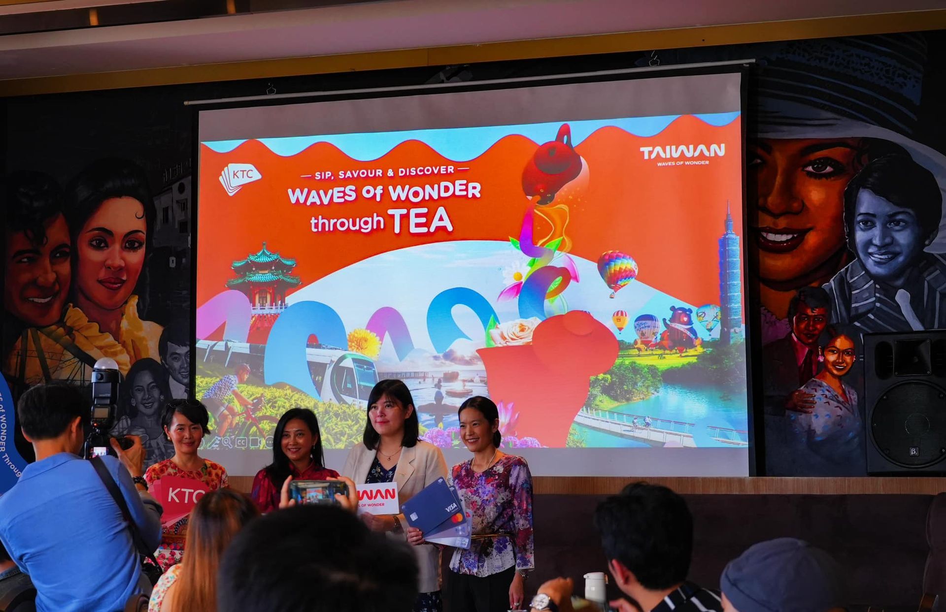 A group of people are standing in front of a large screen that says waves of wonder through tea.