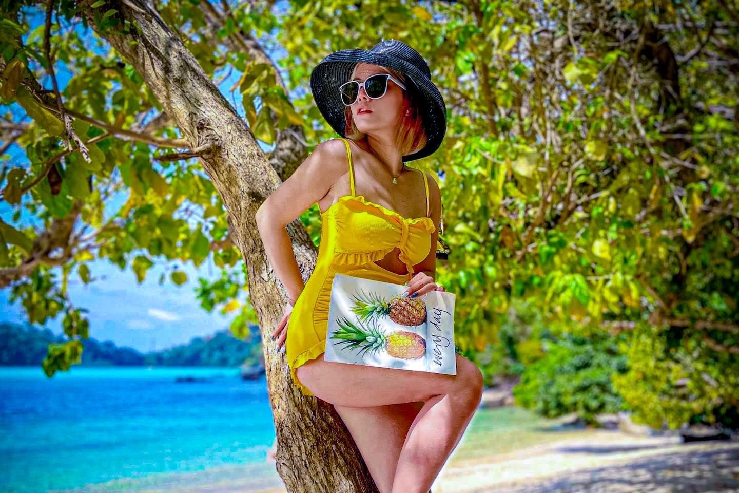 A woman in a yellow dress is leaning against a tree on the beach.