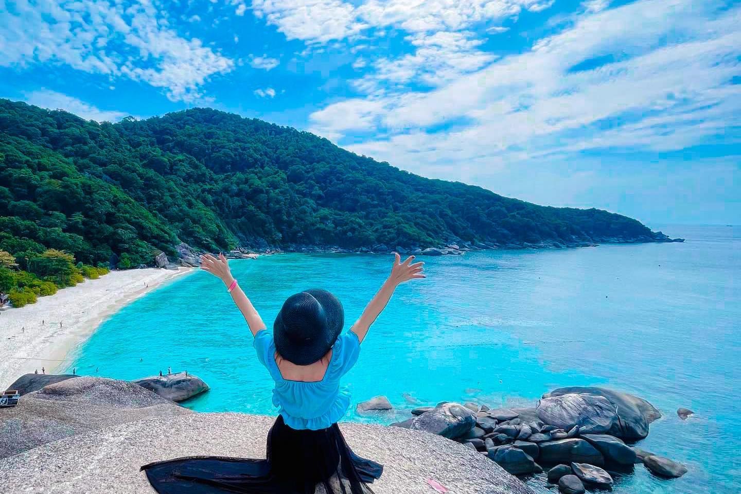 A woman is sitting on a rock overlooking a beach with her arms outstretched.
