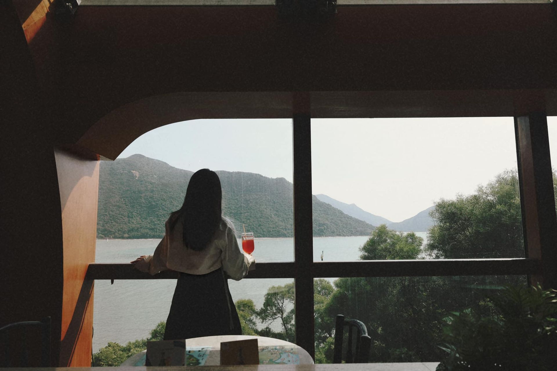 A woman is standing in front of a large window looking out over a lake.