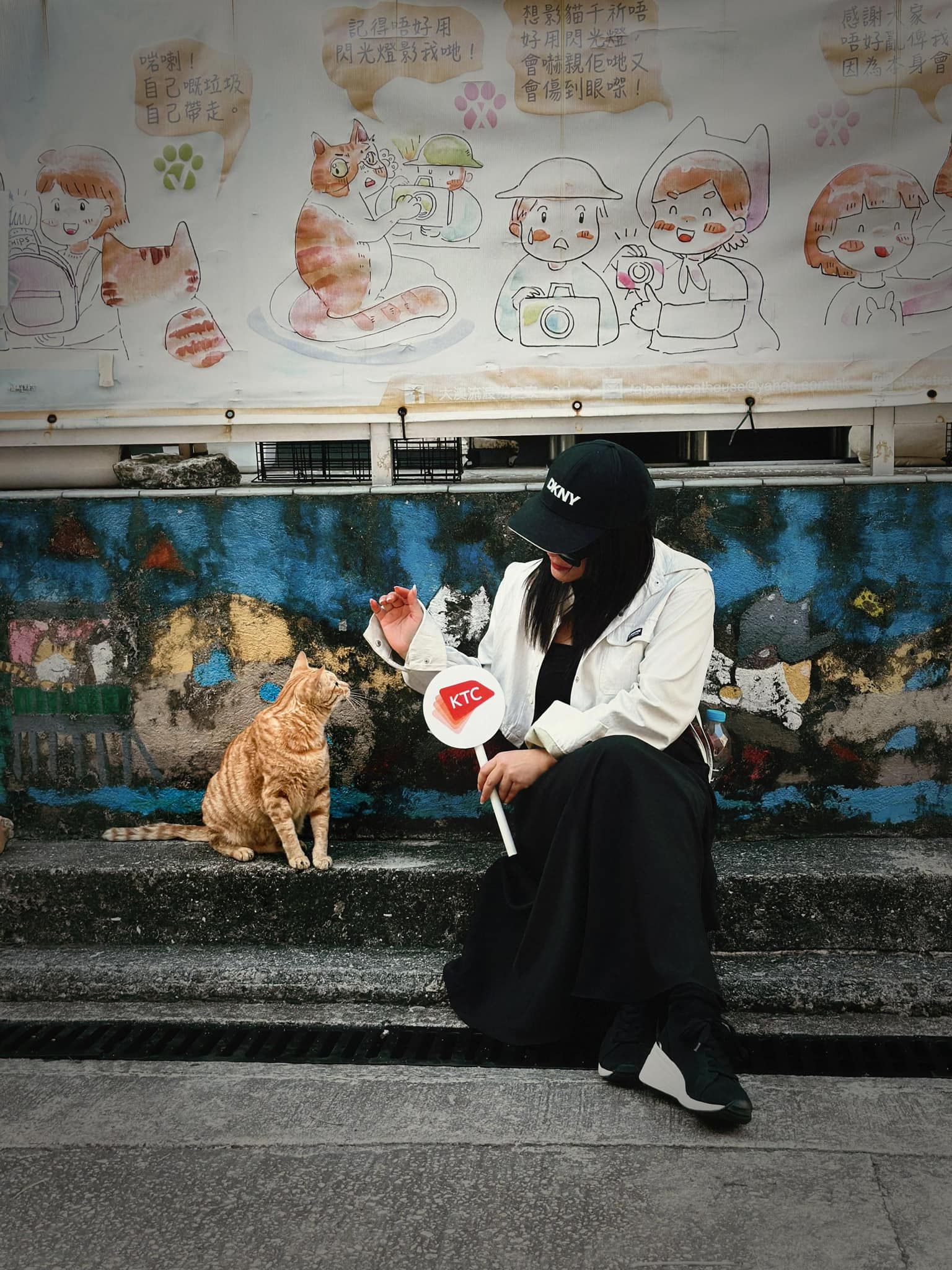 A woman is sitting on the sidewalk next to a cat.