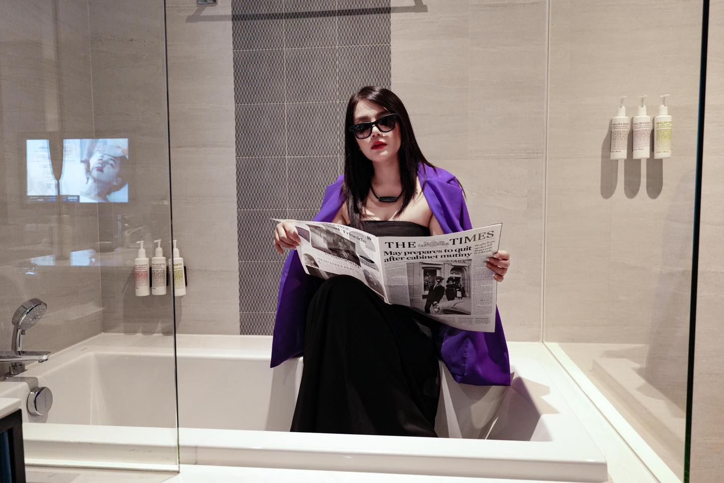 A woman is sitting in a bathtub reading a newspaper.