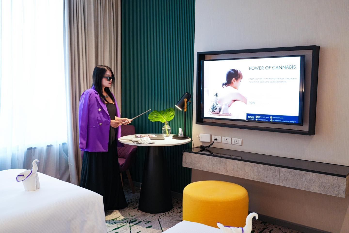 A woman in a purple jacket is standing in a hotel room in front of a television.