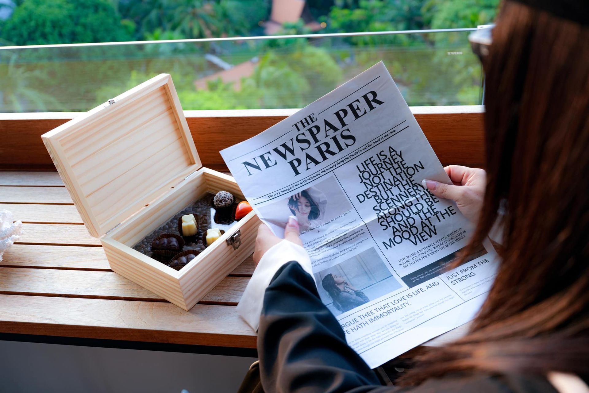 A woman is reading a newspaper next to a box of chocolates.