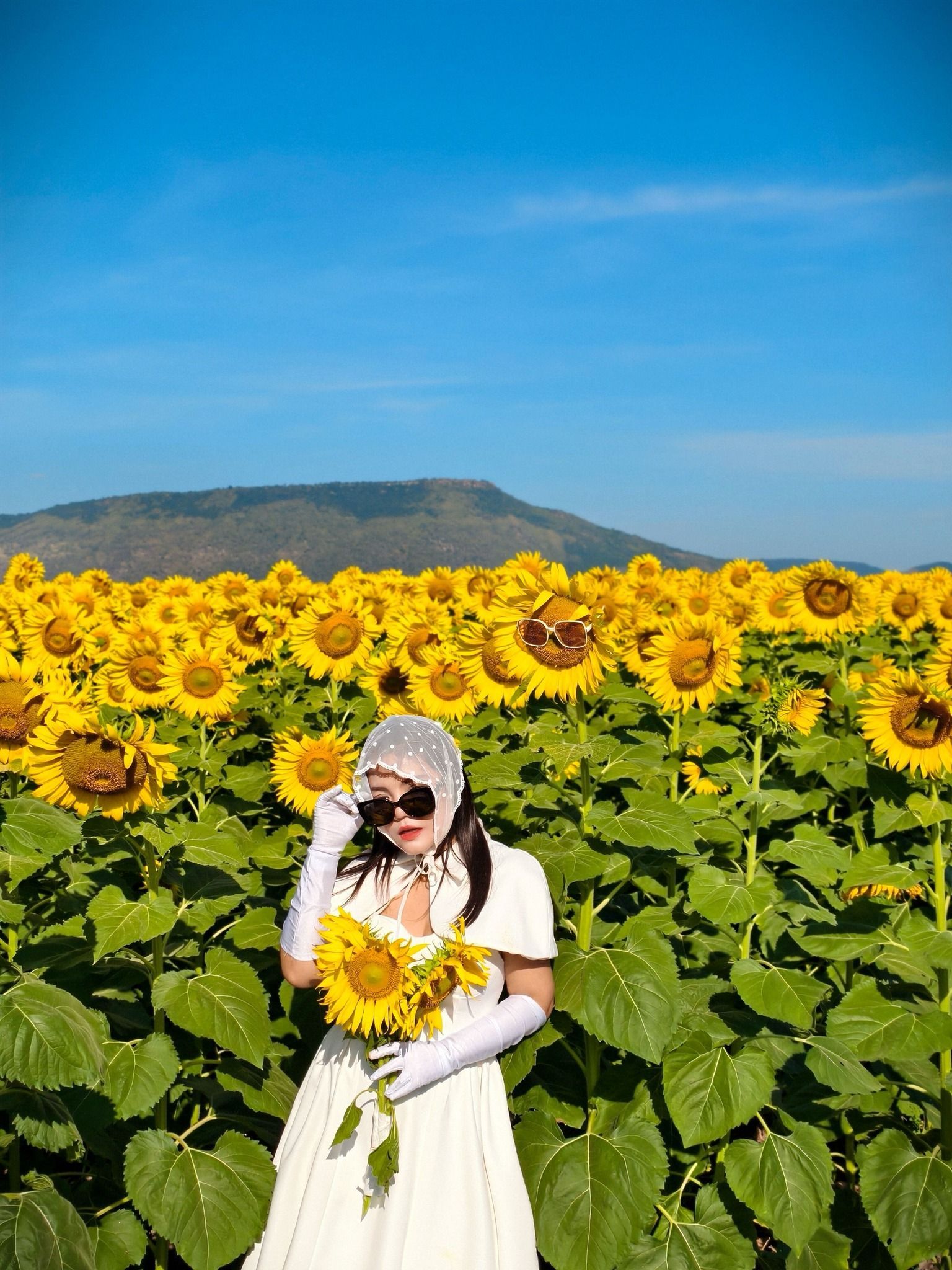 A woman in a white dress is standing in a field of sunflowers holding a bouquet of sunflowers.