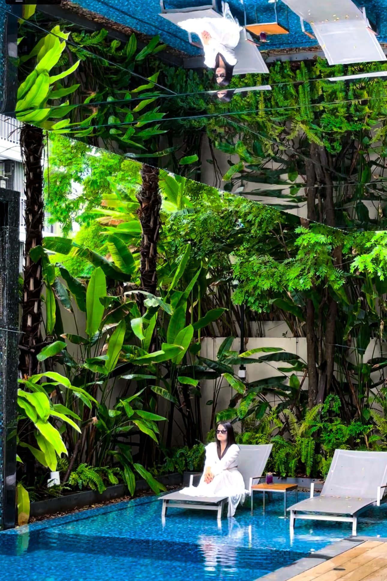 A woman is sitting on a lounge chair next to a swimming pool surrounded by trees.