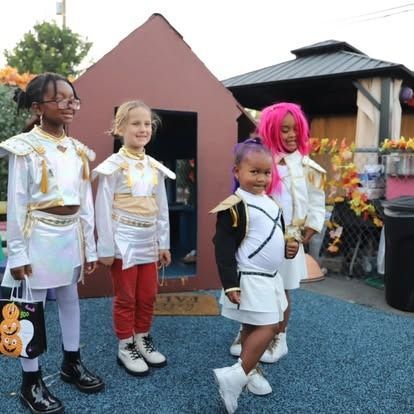 Four children in matching white and gold costumes pose outdoors in front of a small playhouse during Halloween.
