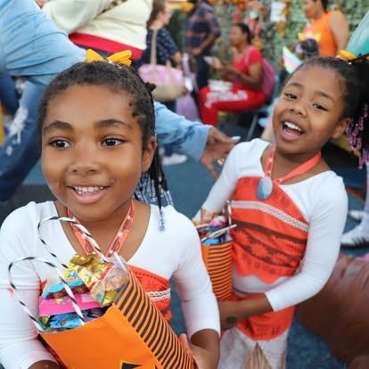 Two children wearing Moana costumes and holding orange trick-or-treat bags smile during a Halloween event.