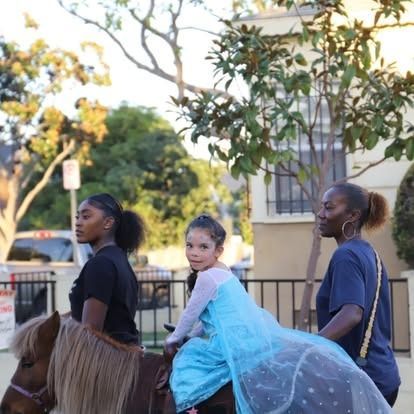 A child in a blue princess dress rides a brown pony outdoors, accompanied by two individuals walking alongside.