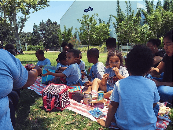 A group of people sitting on a picnic blanket on a sunny day in front of a white building.