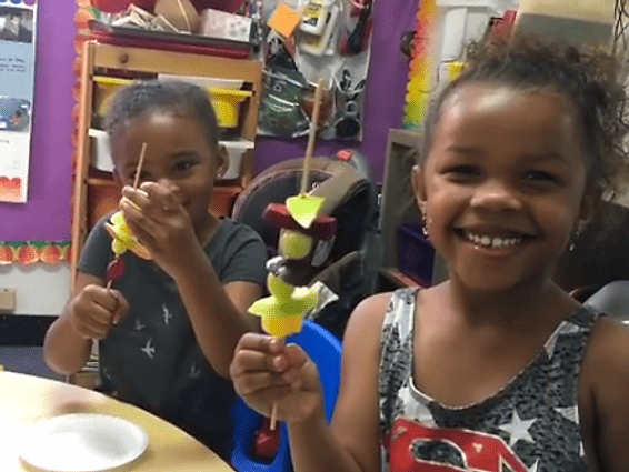 Two children smile as they hold up fruit skewers in a classroom setting.