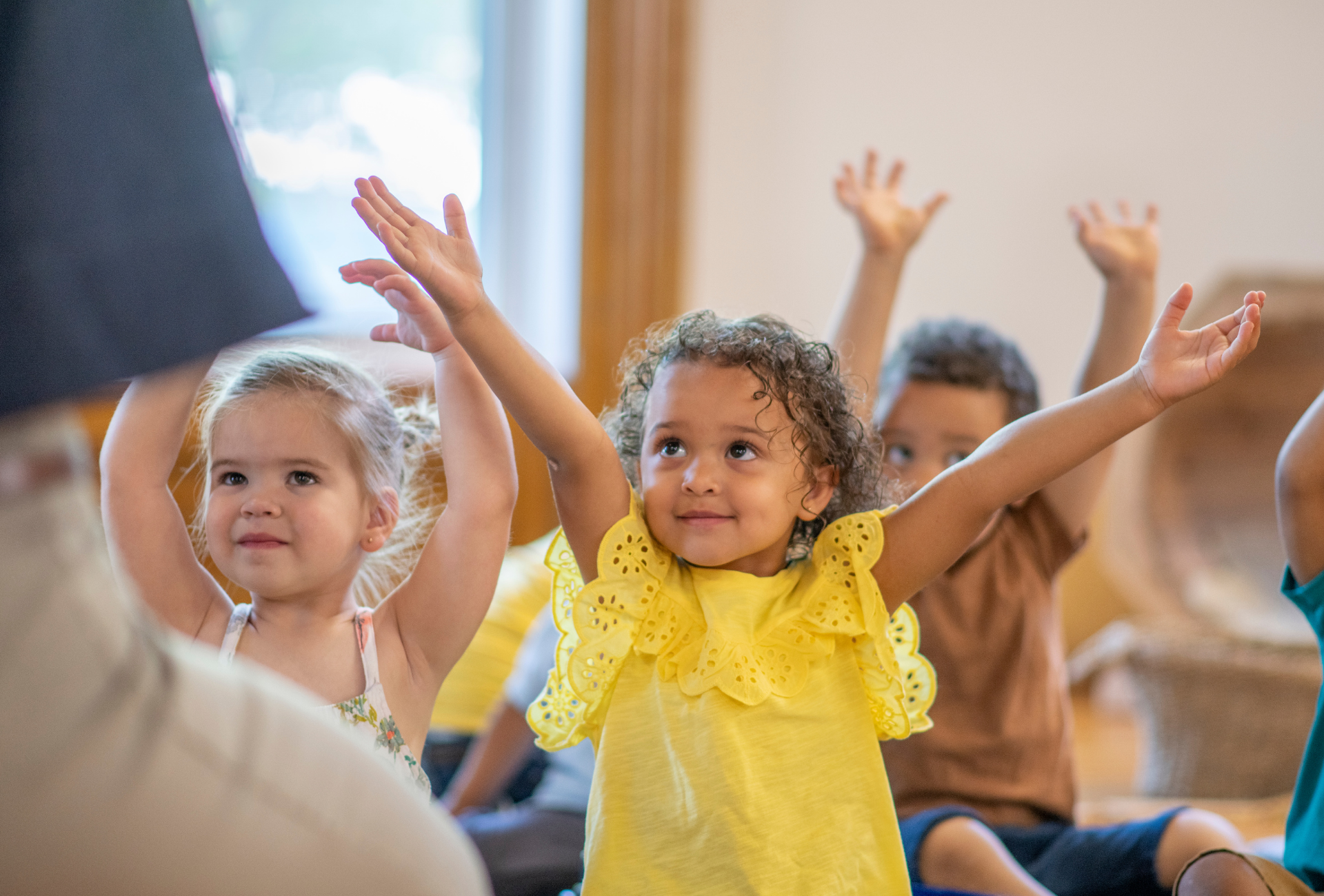Children in a classroom setting raise their hands toward an instructor, smiling with excitement.