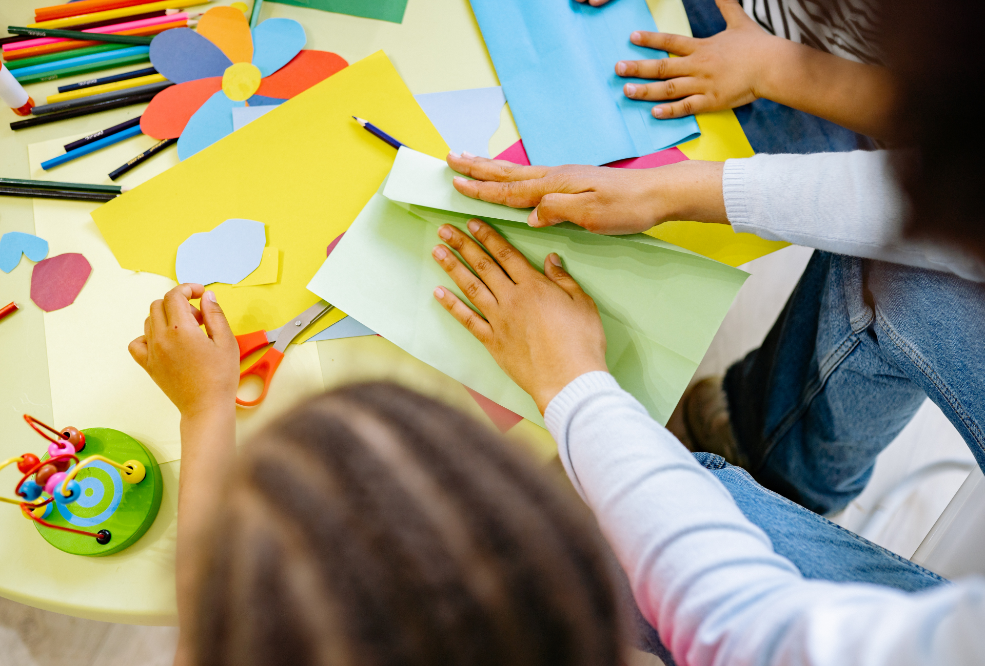 People work together at a table with colorful paper, markers, and craft supplies, folding light green paper.