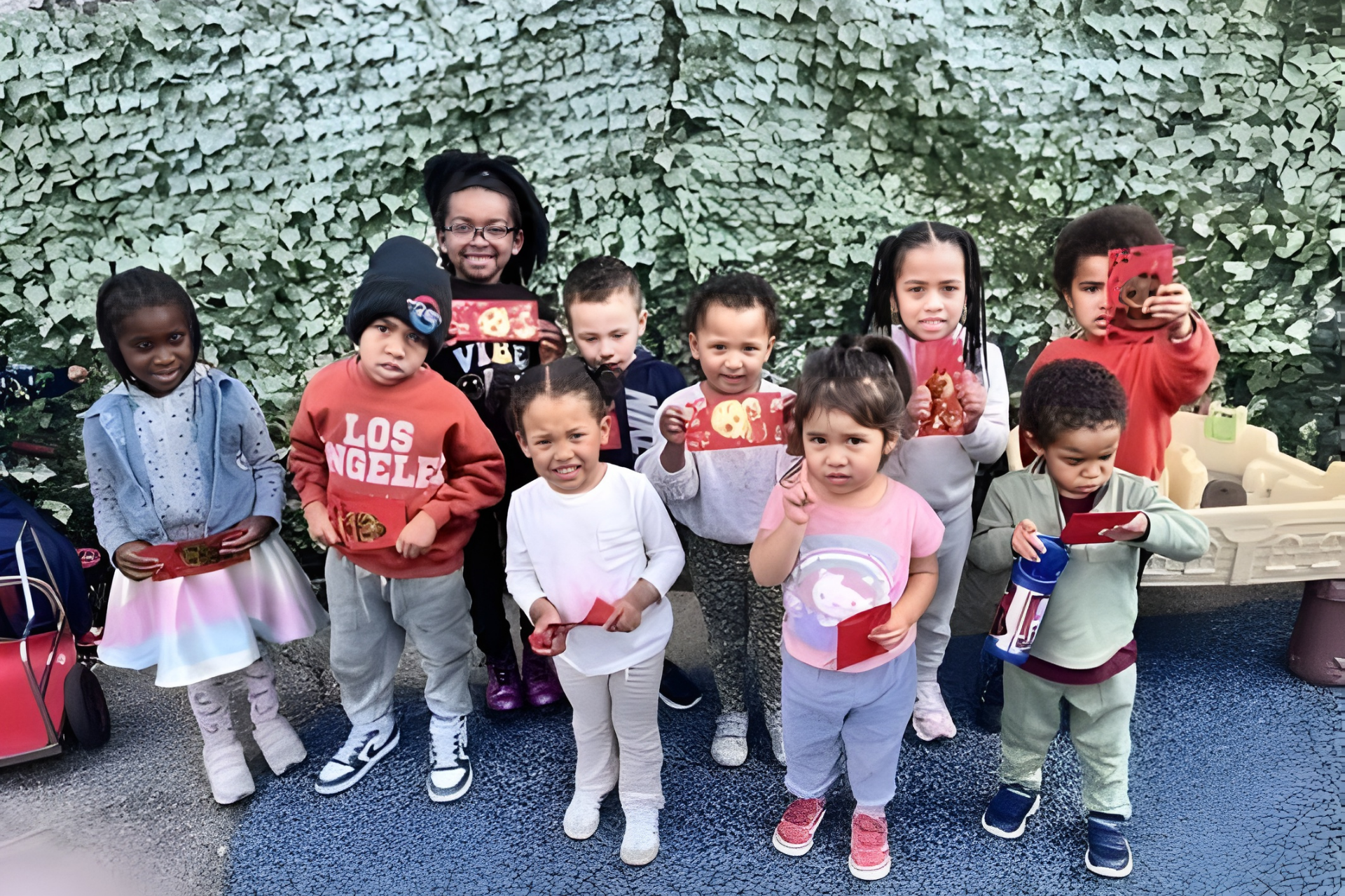 A group of children stand outside against a camouflage-patterned wall, holding small red envelopes.