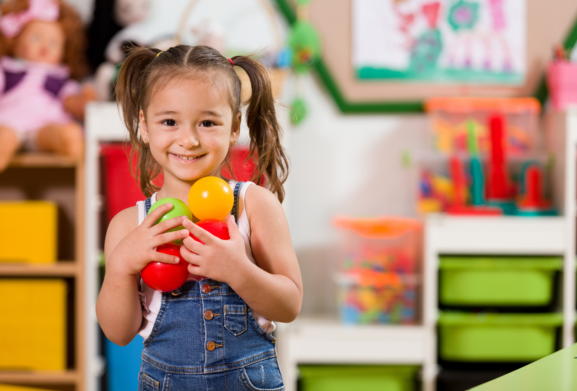A smiling child wearing denim overalls holds three colorful toy balls in a playroom filled with shelves and toy bins.