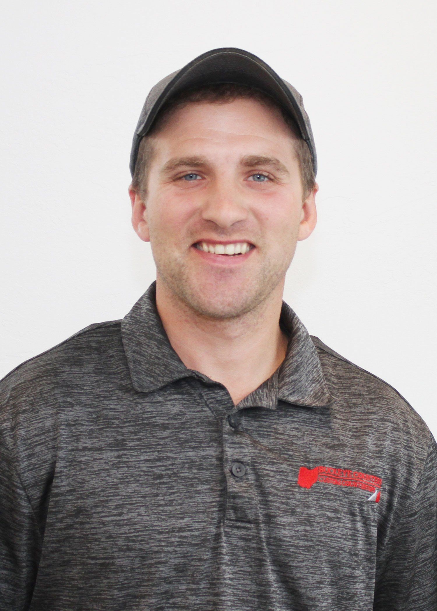 Man smiling, wearing a baseball cap and grey polo shirt with a logo, in front of a white wall.