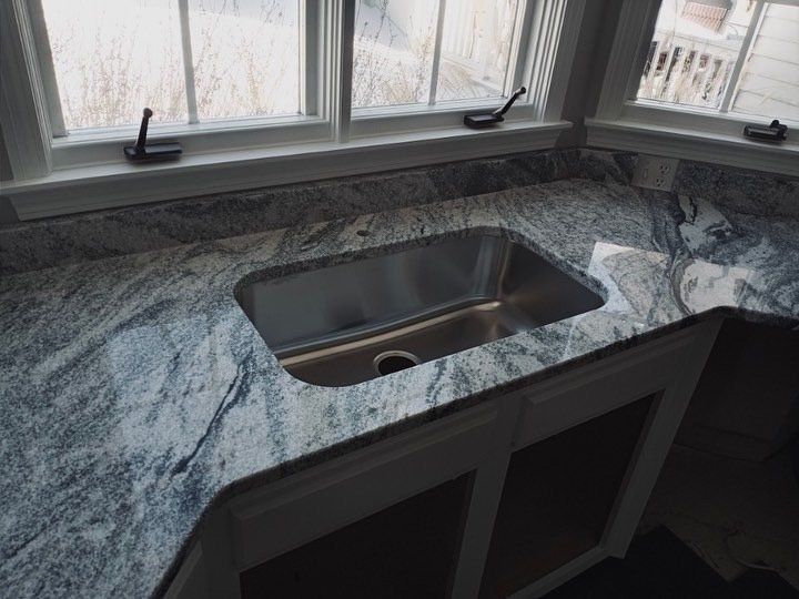 Granite countertop with a stainless steel sink under a window. Cabinets below.