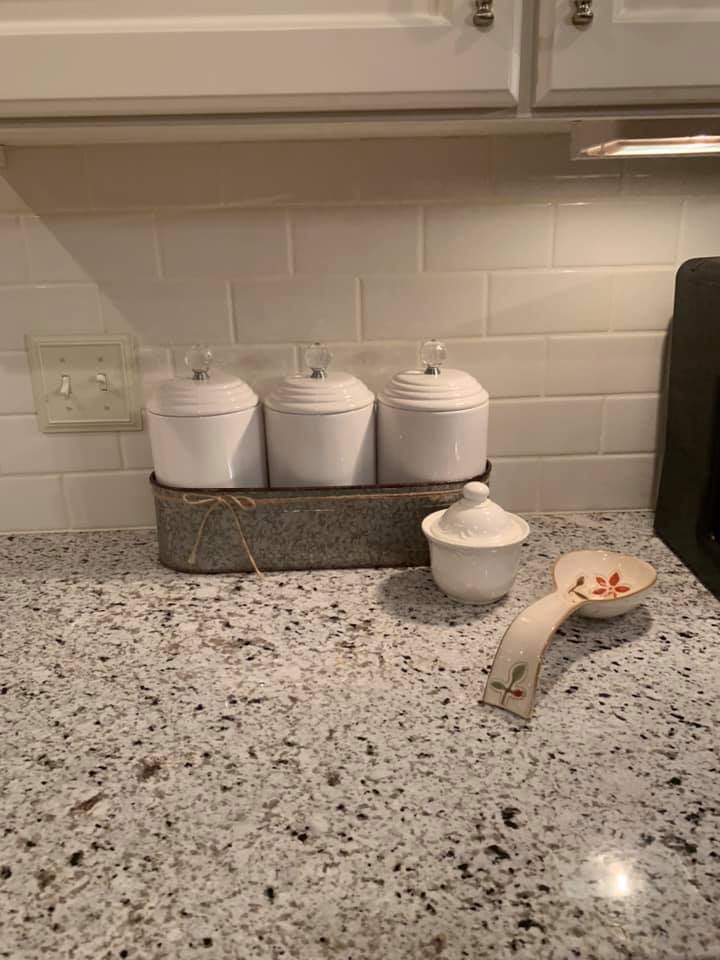 Three white canisters on a speckled countertop, a small covered jar, and a decorative spoon.