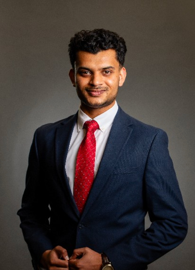 Man in a navy suit and red tie standing against a dark background, smiling