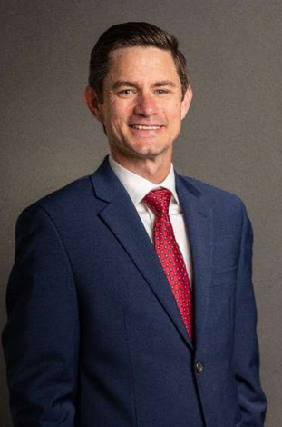 Smiling man in a navy suit, white shirt, and red tie against a gray background